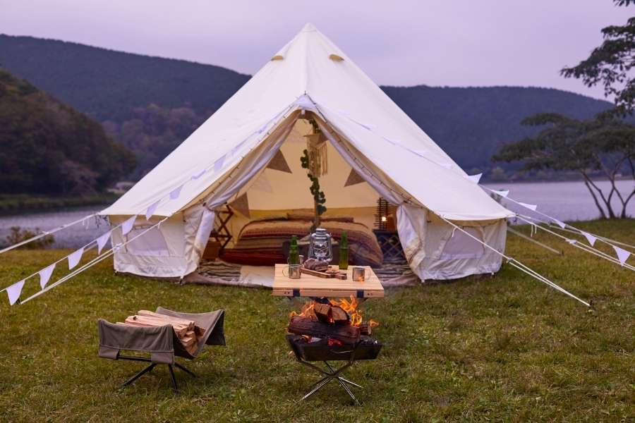 A large white bell tent pitched by a tranquil lake at dusk, designed to create a cozy and relaxing tent experience with a lit fire pit and outdoor seating in the foreground.
