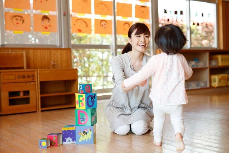A teacher kneeling on the floor, smiling and welcoming a young child who walks towards her next to a tower of alphabet blocks, designed to encourage language development.
